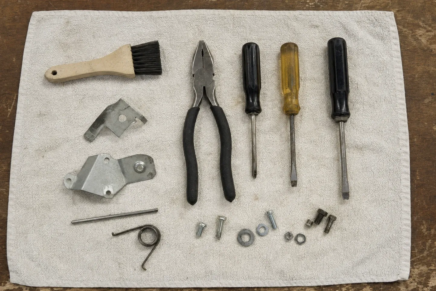 Parts brush, pliers and screwdrivers on a towel beside two hinge brackets, a torsion spring, screws and washers
