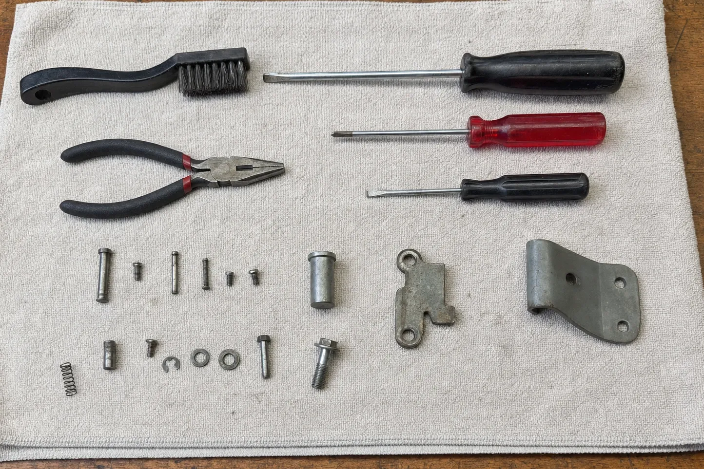 Wire brush, pliers and three screwdrivers laid out on canvas above two rows of pins, screws, washers and two door-hinge brackets